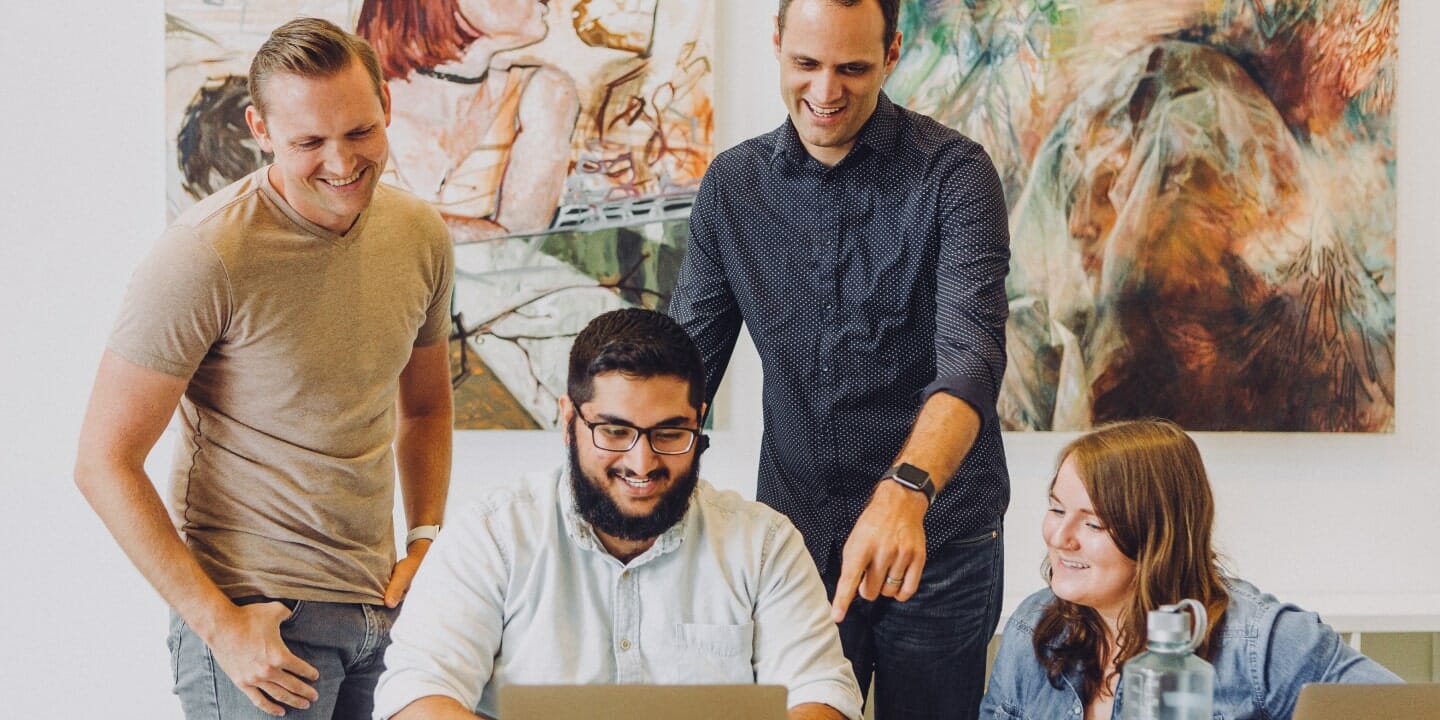 A group of four people, three men and a women, stand and sit around a laptop smiling.